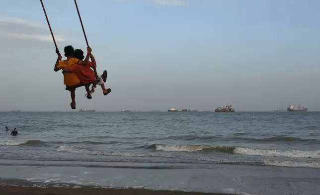 Two children swing together as a mix of bulk carriers, cargo ships, and service vessels line the horizon in the Strait of Hormuz off Bandar Abbas, Iran, Sunday, April 26, 2026.(Razieh Poudat/ISNA via AP)