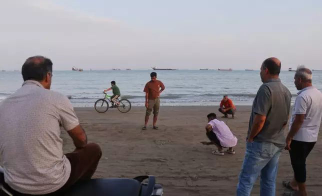 Men gather along the shore, some crouching and watching a game, as a mix of bulk carriers, cargo ships, and service vessels line the horizon in the Strait of Hormuz off Bandar Abbas, Iran, Monday, April 27, 2026.(Razieh Poudat/ISNA via AP)