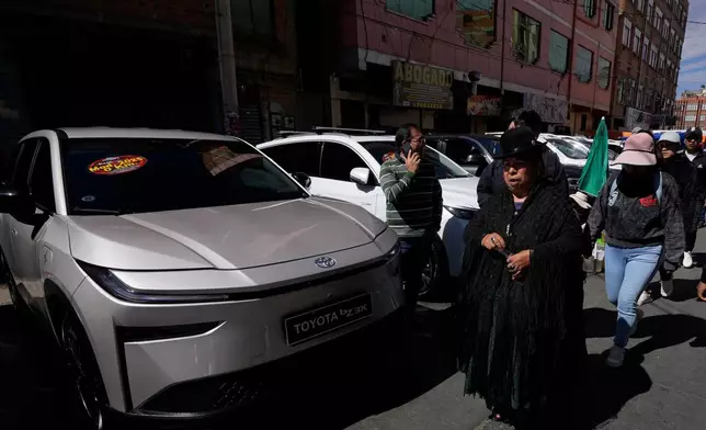 People walks past electric cars displayed for sale at "La Feria de la 16 de Julio," in El Alto, Bolivia, Sunday, April 19, 2026. (AP Photo/Juan Karita)