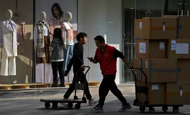 FILE - Workers push carts loaded with goods past an employee who adjusts clothing on the mannequins at a fashion boutique in Beijing, Oct. 13, 2021. (AP Photo/Andy Wong, File)