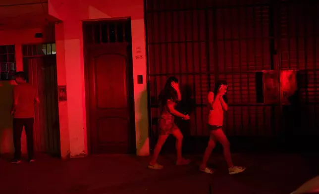 Illuminated by the lights of a police patrol car, neighbors walk past a car repair and auto parts shop targeted in a dynamite attack in Trujillo, Peru, Sunday, May 24, 2026. (AP Photo/Rodrigo Abd)