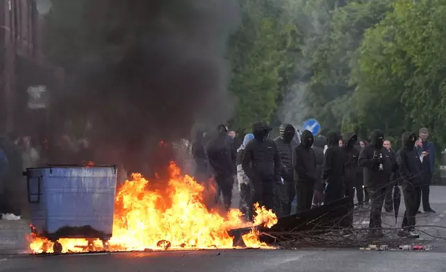 Masked protesters stand by burning trash containers on Ligoniel Road in Belfast, Northern Ireland, on Tuesday, June 9, 2026, after the arrest of a Sudanese man accused of stabbing a man in the northern part of the city. (PA via AP)