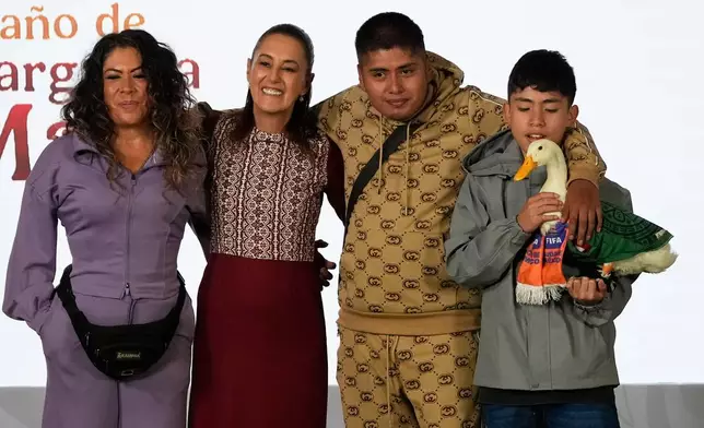 Merlin the duck, dressed in a Mexican national soccer team jersey, attends the daily press conference by Mexican President Claudia Sheinbaum, second from left, along with his caretakers Carla Gomez and her sons Carlos, and Christian who holds Merlin, at the National Palace in Mexico City, Monday, June 22, 2026. (AP Photo/Marco Ugarte)