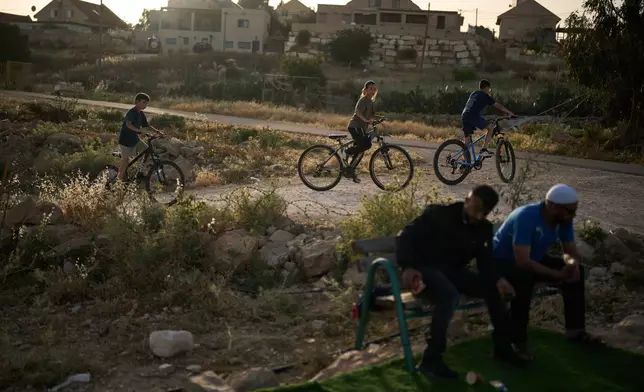 Young Israeli settlers ride bicycles past Palestinian men sitting near a small soccer pitch in the West Bank Bedouin village of Umm al-Khair, Tuesday, May 12, 2026. (AP Photo/Leo Correa)
