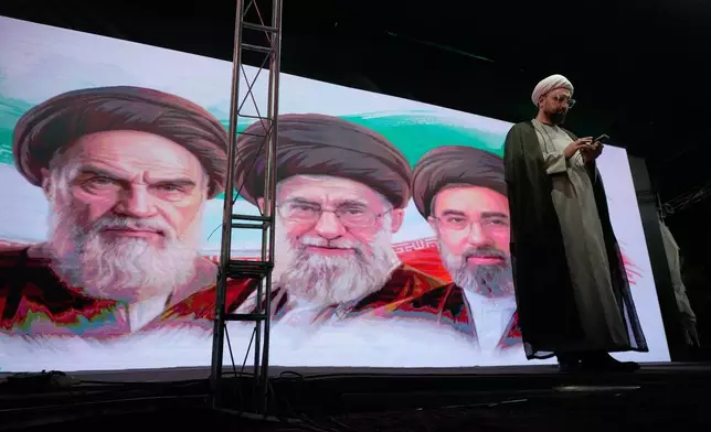 A cleric checks his cell phone on stage in front of a screen displaying portraits of the late Iranian revolutionary founder Ayatollah Khomeini, left, late Supreme Leader Ayatollah Ali Khamenei, center, and Supreme Leader Ayatollah Mojtaba Khamenei, during a pro-government gathering in Tehran, Iran, Sunday, June 7, 2026. (AP Photo/Vahid Salemi)