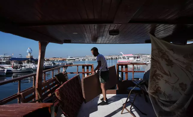 With the statue of Our Lady of the Seas visible in the background, Ali Bazzi, 31, who was displaced from his home in the village of Toura during the war between Israel and Hezbollah, arranges cushions aboard a small boat where he has been living in the southern port city of Tyre, Lebanon, Saturday, June 20, 2026. (AP Photo/Hassan Ammar)