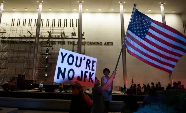 Protestors wave a U.S. and signs as workers prepare to remove President Donald Trump's name from the John F. Kennedy Center for the Performing Arts in Washington, Saturday, June 13, 2026. (AP Photo/Cliff Owen)