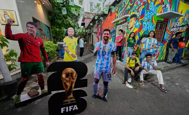 Enthusiasts sit beside wall graffiti as cutouts of Portugal's Cristiano Ronaldo, Brazil's Neymar and Argentina's Lionel Messi are displayed on a street on the occasion of World Cup soccer tournament, Thursday, June 18, 2026, in Kolkata, India. (AP Photo/Bikas Das)