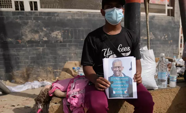 Kevin Tovar sits near his damaged home holding leaflets with a photo of his missing father in La Guaira, Venezuela, Friday, June 26, 2026, two days after twin earthquakes struck the country. (AP Photo/Ariana Cubillos)