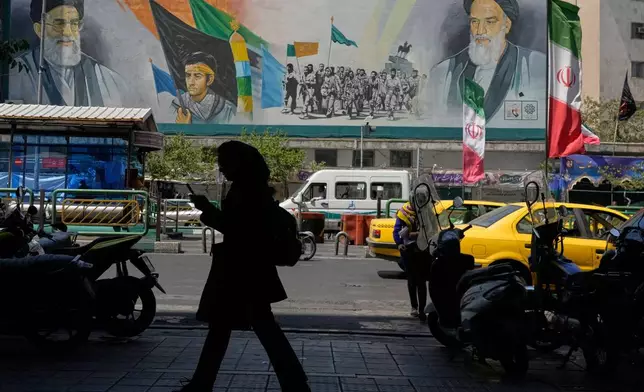 A woman walks on a sidewalk in front of a mural showing portraits of the late Iranian revolutionary founder Ayatollah Khomeini, right, the slain Supreme Leader Ayatollah Ali Khamenei, who was killed in U.S. and Israeli strikes on Feb. 28, and Basij paramilitary forces, at the Islamic Revolution street in Tehran, Iran, Monday, June 8, 2026. (AP Photo/Vahid Salemi)