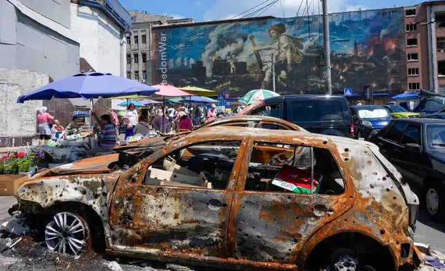 People buy food at an improvised outdoor market, burnt cars in the foreground, surrounded by damaged buildings covered with street artists paintings close to a big city marketplace that was ruined recently by Russian missiles in Kyiv, Ukraine, Monday, June 22, 2026. (AP Photo/Efrem Lukatsky)