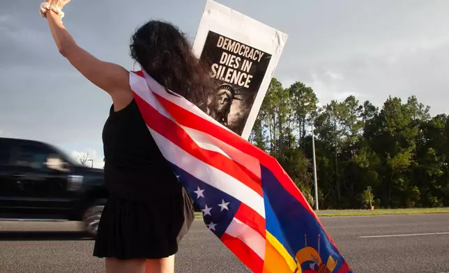 Patricia Ramos, of Palm Coast, wears a flag and waves at passing cars during a vigil for a Mexican man who was struck and killed by a tractor trailer while running from an encounter with Immigration and Customs Enforcements officers the day before, Wednesday, July 15, 2026, in St. Augustine, Fla. (AP Photo/Kelly Jordan)