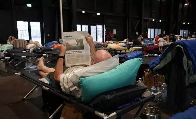 An evacuee reads a newspaper report about wildfires in southwest France as he lays on a camp bed at a temporary shelter in Bordeaux, France, Tuesday, July 28, 2026. (AP Photo/Eva van Dam)