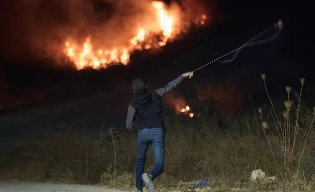 A Palestinian uses a slingshot during clashes with Israeli settlers as brush burns after it was set ablaze to prevent them from approaching the West Bank village of Sinjil, Friday, July 17, 2026. (AP Photo/Leo Correa)