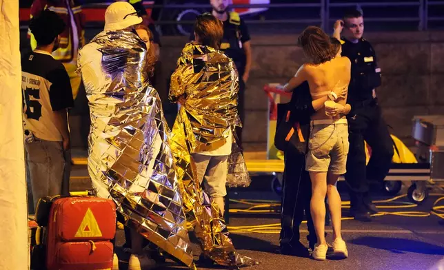 Two people embrace near others wearing thermal blankets as they stand near emergency personnel in Berlin, early Sunday, July 26, 2026, following the cancellation of Christopher Street Day festivities. (AP Photo/Markus Schreiber)