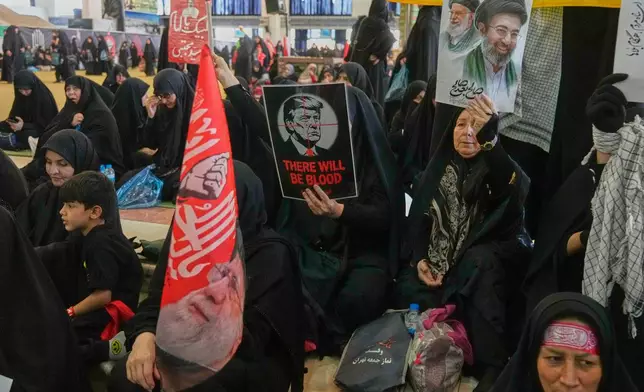 A woman holds a poster depicting U.S. President Donald Trump in the crosshairs of a rifle scope with the English words "There Will Be Blood," while another woman holds a portrait of the late Supreme Leader Ali Khamenei and his son, current Supreme Leader Mojtaba Khamenei, as mourners gather to commemorate the late leader at the Imam Khomeini Mosalla Grand Mosque in Tehran, Iran, Tuesday, July 14, 2026. (AP Photo/Vahid Salemi)