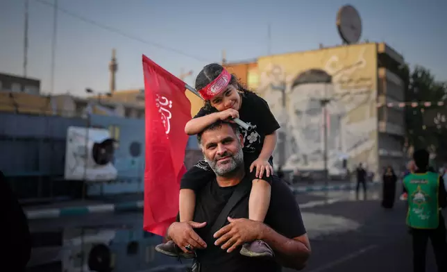 A man carries a child holding a red Shiite religious flag outside the Imam Khomeini Mosalla Grand Mosque during the funeral ceremonies for slain Iranian Supreme Leader Ayatollah Ali Khamenei and members of his family in Tehran, Iran, Saturday, July 4, 2026. (AP Photo/Altaf Qadri)
