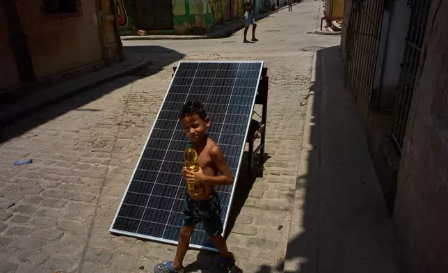 A child walks with a bottle of oil past a solar panel set up on the street to charge batteries during a blackout in Havana, Cuba, Monday, July 6, 2026. (AP Photo/Ramon Espinosa)