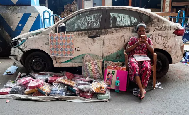 With a display of handbags, a roadside vendor scrolls on her mobile, awaits for customers in front of a parked vehicle, in Kolkata, India, Saturday, July 18, 2026. (AP Photo/Bikas Das)