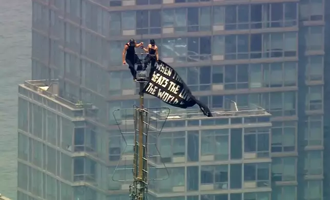 Two people stand on the tip of the antenna of the Empire State Building in New York while holding a banner on Wednesday, July 1, 2026. (WABC-TV via AP)