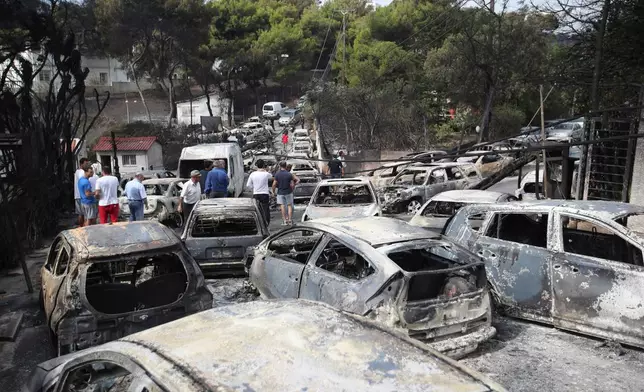FILE - People stand amid the charred remains of burned-out cars following a deadly wildfire, in Mati east of Athens, Tuesday, July 24, 2018. (AP Photo/Thanassis Stavrakis, File)