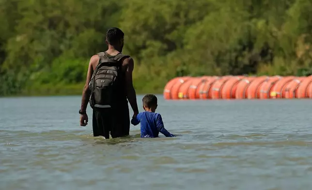 FILE - Migrants walk past large buoys being used as a floating border barrier on the Rio Grande, Aug. 1, 2023, in Eagle Pass, Texas. (AP Photo/Eric Gay, File)