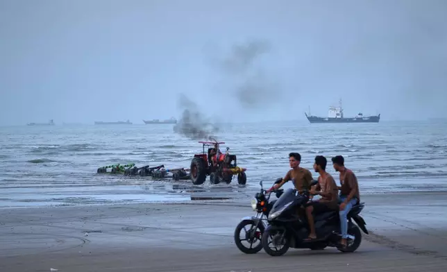 Young men ride motorcycles along the shore as a tractor pulls equipment from the water and commercial vessels appear anchored in the Strait of Hormuz off Bandar Abbas, Iran, Monday, July 27, 2026. (Razieh Poudat/ISNA via AP)