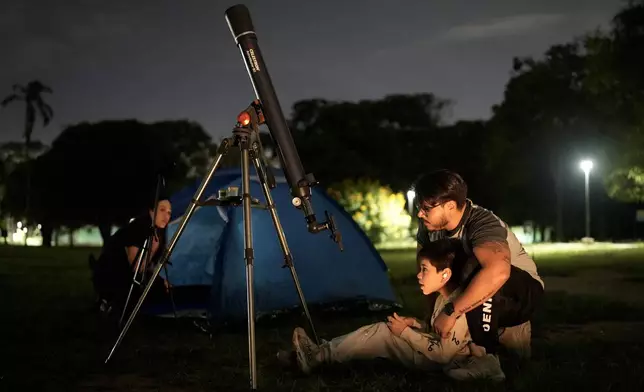 Hector Garcia looks at the moon through a telescope and a cellphone adapted with his son during a lunar eclipse in Caracas, Venezuela, Thursday, Aug. 27, 2026. (AP Photo/Ariana Cubillos)