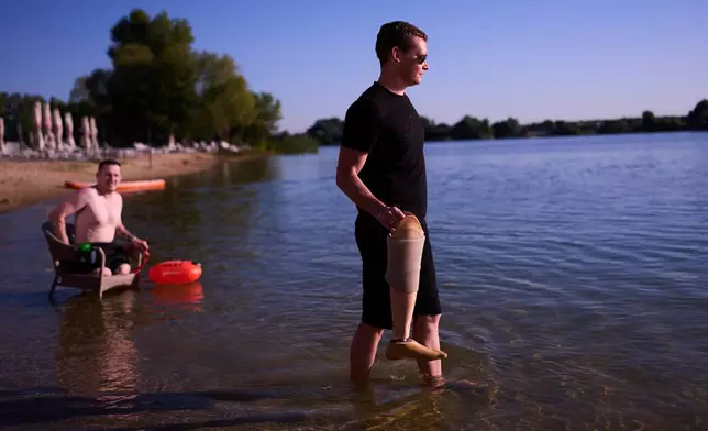 A man holds the prosthetic leg of a Ukrainian war veteran as Maxim Yuzvyak, left, also a veteran, sits on a chair during a sporting event to encourage people and injured soldiers to take part in sports at a lake on the outskirts of Kyiv, Monday, Aug. 24, 2026, during the 35th anniversary of Ukraine's Independence Day. (AP Photo/Francisco Seco)