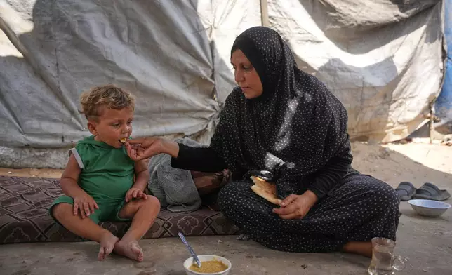 A Palestinian woman feeds a child a meal cooked using combustible materials collected from a garbage dump amid a shortage of cooking gas in Deir al-Balah, central Gaza Strip, Saturday, Aug. 22, 2026. (AP Photo/Abdel Kareem Hana)