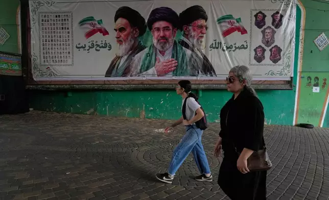Women walk past a banner showing portraits of the late Iranian revolutionary founder Ayatollah Khomeini, left, Supreme Leader Ayatollah Mojtaba Khamenei, center, and the late Supreme Leader Ayatollah Ali Khamenei, who was killed in the U.S. and Israel strike on Feb. 28, under a bridge in northern Tehran, Iran, Sunday, Aug. 2, 2026. (AP Photo/Vahid Salemi)