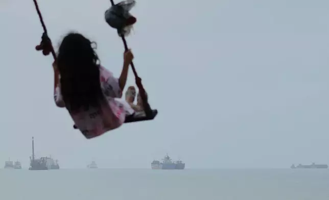 A girl swings along the shore as cargo ships and other commercial vessels appear anchored in the Strait of Hormuz off Bandar Abbas, Iran, Monday, July 27, 2026. (Razieh Poudat/ISNA via AP)