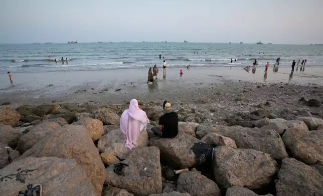 Two people sit on rocks overlooking a beach as others swim and commercial vessels lie anchored in the Strait of Hormuz off Bandar Abbas, Iran, Friday, Sept. 4, 2026. (AP Photo/Vahid Salemi)