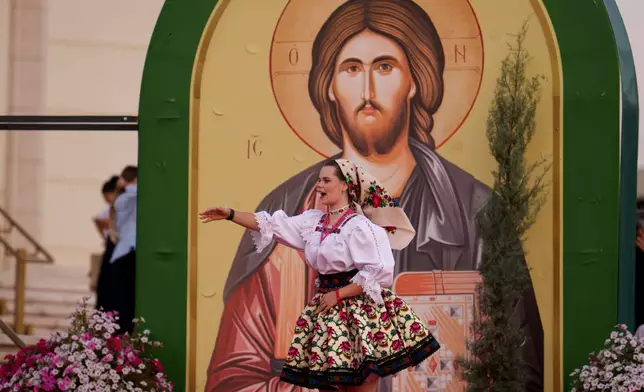 A young woman wearing a traditional outfit gestures backdropped by a religious icon before the Procession of Lights, part of a four-day program of the International Meeting of Orthodox Youth (ITO), in Bucharest, Romania, Wednesday, Sept. 2, 2026. (AP Photo/Andreea Alexandru)