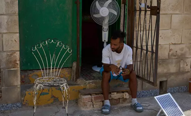 A man charges his fan's battery at his doorway using a small solar panel set up on the street during a blackout in Havana, Cuba, Friday, Sept. 18, 2026. (AP Photo/Ramon Espinosa)