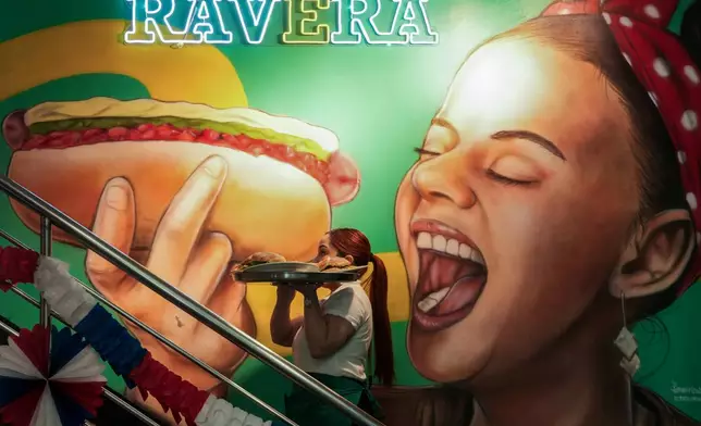 A waitress carries a tray with "completos", traditional Chilean hot dogs topped with mashed avocado, diced tomatoes and mayonnaise, past a mural dedicated to the dish at the Ravera restaurant in downtown Santiago, Chile, Thursday, Sept. 10, 2026. (AP Photo/Esteban Felix)