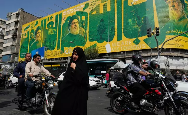 A woman walks and vehicles drive in an intersection in front of a billboard showing portraits of the late Iranian Supreme Leader Ayatollah Ali Khamenei, right, late leaders of Lebanon's Hezbollah group Hassan Nasrallah, center left, and Hashem Safieddine, left, marking the second anniversary of the killing of Hassan Nasrallah, in Tehran, Iran, Sunday, Sept. 27, 2026. (AP Photo/Vahid Salemi)