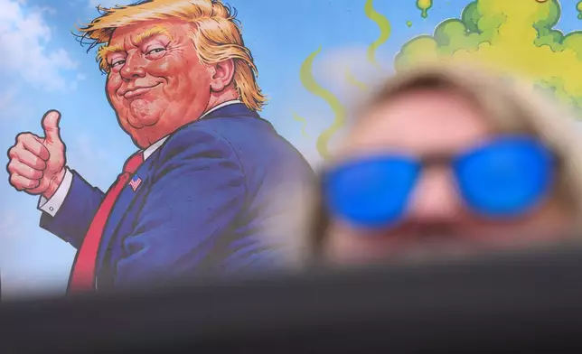 A protester wears sunglasses and sits in front of a poster depicting U.S. President Donald Trump during his visit to the Irish Open at the Trump International Golf Links course in Doonbeg, Ireland, Sunday, Sept. 13, 2026.(AP Photo/Kin Cheung)