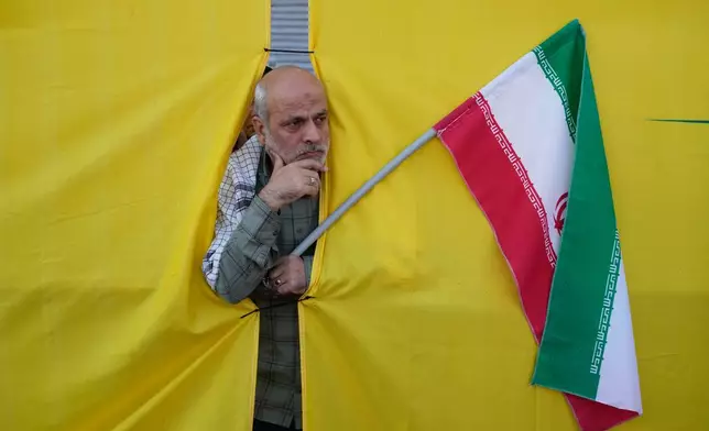 A man holding an Iranian flag peers through an opening in a curtain during the state-organized “Janfada,” or “Sacrifice for Iran,” military-style parade in Tehran, Iran, Friday, Sept. 18, 2026. (AP Photo/Vahid Salemi)