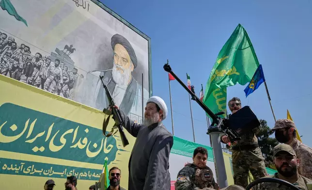 An armed Shiite cleric rides aboard a military vehicle past a billboard depicting Ayatollah Ruhollah Khomeini, founder of the Islamic Republic, during the state-organized "Janfada," or "Sacrifice for Iran," military-style parade in Tehran, Iran, Friday, Sept. 18, 2026. (AP Photo/Vahid Salemi)