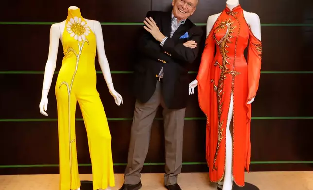 FILE - Fashion and Costume Designer Bob Mackie poses with two of his iconic designs, a scarlet red satin gown worn by Cher in 1975, right, and a marigold jersey jumpsuit worn by Cher between 1971-1976, left, in London on Aug. 16, 2018. (AP Photo/Kirsty Wigglesworth, File)