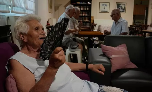 FILE - Jenny Sakellariou uses a hand fan while men play backgammon in an air-conditioned cooling center operated by the Athens municipality throughout a heat wave in Athens, Greece, Aug. 24, 2026. (AP Photo/Thanassis Stavrakis, File)