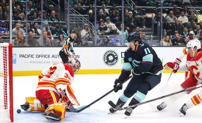 Seattle Kraken right wing Mackie Samoskevich (11) gets the puck past Calgary Flames goaltender Dustin Wolf (32) as defenseman Zach Whitecloud, right, defends during the first period of an NHL hockey game Sunday, Oct. 4, 2026, in Seattle. (AP Photo/Jason Redmond)