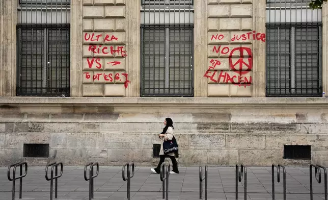 A woman walks past a building with graffiti that reads "The ultra-rich against everyone" and "No justice, no peace. RIP El Hacen" after the weekend protests over the cost of living turned into rioting and looting across France, in Paris, Wednesday, Oct. 21, 2026. (AP Photo/Teresa Suarez)