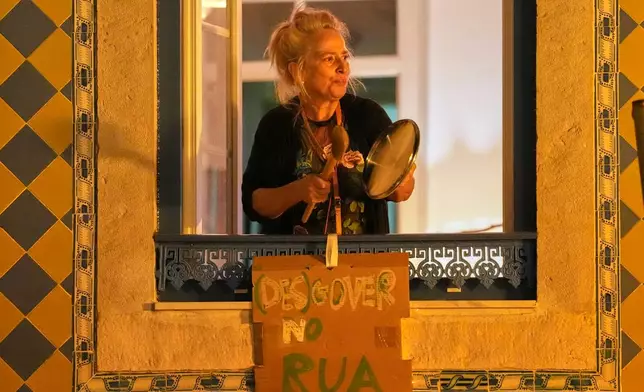A woman protests from a window of her home as demonstrators gather to demand affordable housing outside Portugal's Parliament in Lisbon, Tuesday, Oct. 27, 2026. The sign reads in Portuguese: “Out with the (no)government.” (AP Photo/Armando Franca)