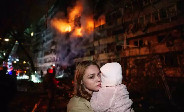 Victoria Deineko holds her daughter Mia while standing in front of a multi-story residential building damaged in a Russian missile strike in Kyiv, Ukraine, Tuesday, Oct. 13, 2026. (AP Photo/Evgeniy Maloletka)