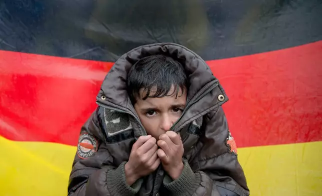 FILE - In this Wednesday, March 9, 2016 file photo a child tries to warm his hands, backdropped by Germany's flag as protesting migrants stage a sit in protest on the railway tracks at northern Greek border station of Idomeni. (AP Photo/Vadim Ghirda, File)