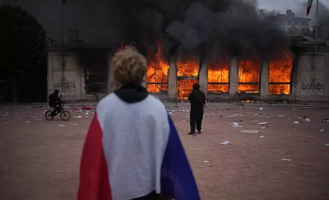A protester wraps a French flag around them in front of the burning tourism office during a demonstration against rising living costs and in solidarity with students’ grievances in Lyon, France, Saturday, Oct. 17, 2026. (AP Photo/Laurent Cipriani)