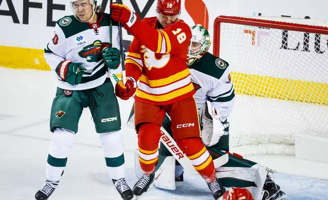 Minnesota Wild's Bobby Brink, left, is hit by the puck as he battles with Calgary Flames' Ben Jones, center, as Wild goalie Jesper Wallstedt, right, reactss during the second period of an NHL hockey game in Calgary, Alberta, Thursday, Oct. 22, 2026. (Jeff McIntosh/The Canadian Press via AP)