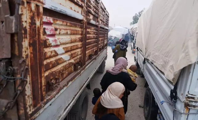 Women carry young children between trucks loaded with belongings as displaced families prepare to leave al-Talayi camp in Hassakeh, Syria, Tuesday, Oct. 13, 2026. Around 450 families are returning to the Ras al-Ayn countryside, seven years after a Turkish military offensive forced them to flee. (AP Photo/Baderkhan Ahmad)